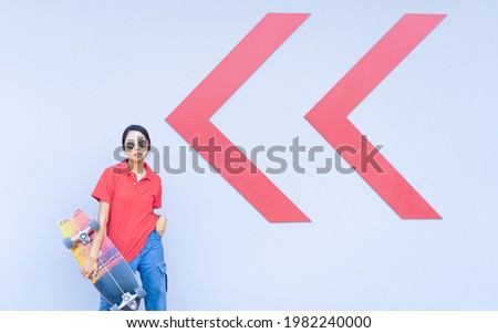 Asian cute girl or woman stand, hold skateboard, looking straight, and act smart at cool wall with arrow in summer holiday.