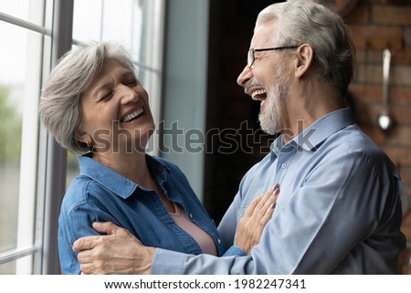 Middle-aged untroubled couple happy homeowners hugging laughing while standing in kitchen at home. Medical insurance cover, dental clinic services for older citizen, carefree midlife and love concept Royalty-Free Stock Photo #1982247341