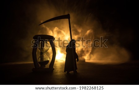 Time concept. Scary view of grim reaper silhouette standing at hourglass with smoke and lights on a dark background. Surreal decorated picture
