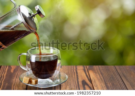closeup coffee pouring from french press into glass cup on wooden table with green blurred background
