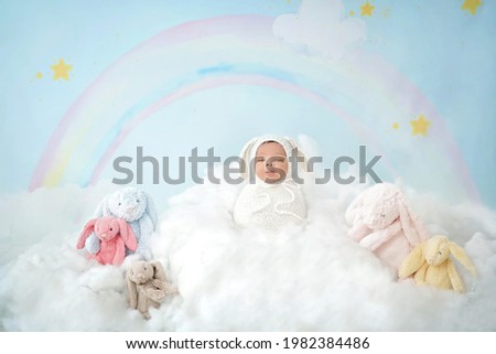 Baby newborn wearing white swaddling wrap standing on white cloud. Newborn is between five rabbit dolls with rainbow and stars on sky blue background.