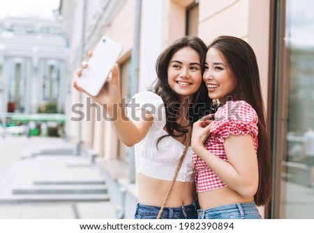 Happy smiling beautiful brunette young women friends in summer clothes taking selfie on mobile phone at summer city street