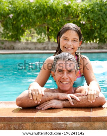 Family portrait of a father and his daughter enjoying the swimming pool together leaning on the stone edge and smiling at the camera during a summer holiday outdoors. Active family lifestyle.