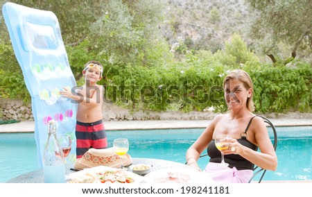 Portrait of a mother and son enjoying fun and healthy food, drinking and eating outdoors in a holiday home garden with a swimming pool during a summer vacation, smiling together. Recreation lifestyle.