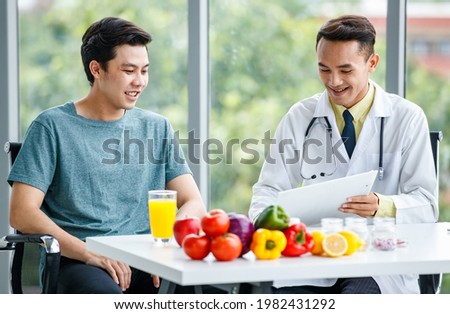 Positive Asian doctor smiling and demonstrating table to male patient while sitting at desk with healthy food in clinic