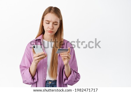 Sad young woman looking at her credit card, holding mobile phone, frowning and sulking with regret, standing against white background