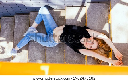 A young woman is lying on the steps. Bright sunlight and shadows fall on the girl. A warm summer day. The girl is wearing jeans and a black men's T-shirt. Creative photography.