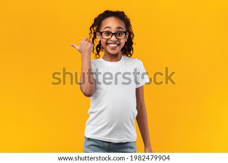 Great Offer. Portrait Of Happy Smiling African American Girl In Glasses Pointing Thumb Aside At Free Blank Space For Text Or Advert Standing Isolated On Yellow Studio Background. Look There, Banner