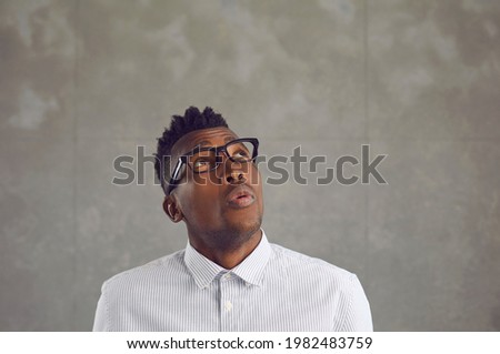 Wow that's interesting. African man sees something curious. Young black guy in white shirt and stylish eyeglasses looks up and whistles surprised by unexpected news on gray copy space background above Royalty-Free Stock Photo #1982483759