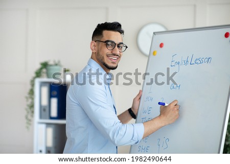 Young Arab male teacher giving online class, explaining new material, writing English rules on blackboard at home office. Remote tutoring and distance education during coronavirus concept