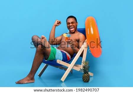 Full length of excited black man drinking tropical cocktail in lounge chair, gesturing YES over blue background. Young African American guy chilling on beach during summer vacation