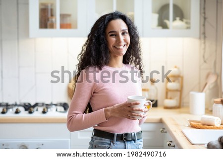 Portrait Of Beautiful Smiling Brunette Woman With Cup Of Coffee In Hands Standing In Kitchen Interior And SLooking At Camera, Happy Young Caucasian Lady Enjoying Hot Drink And Domestic Leisure