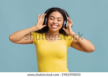 Overjoyed black woman in wireless headset listening to music with closed eyes, enjoying good sound with modern headphones, blue studio background. Rest and relax concept