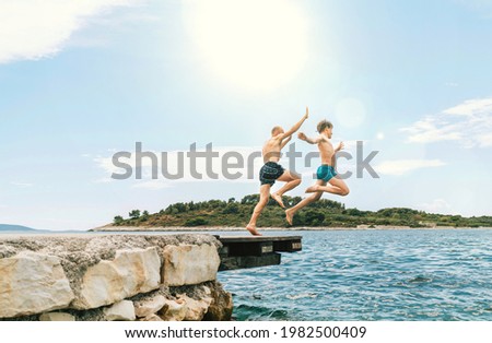 Son with Father having a fun on a merry vacation days on Adriatic sea coast. They dressed swimming trunks jumping to the waives from the boat pier. Family time, fatherhood or childhood concept image. Royalty-Free Stock Photo #1982500409