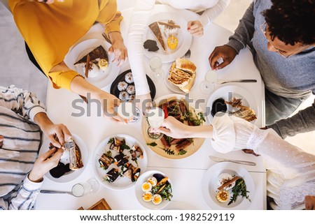 Top view of five multicultural friends sitting at dinning table and having healthy lunch. Women are toasting with fresh lemonade while men eating food.
