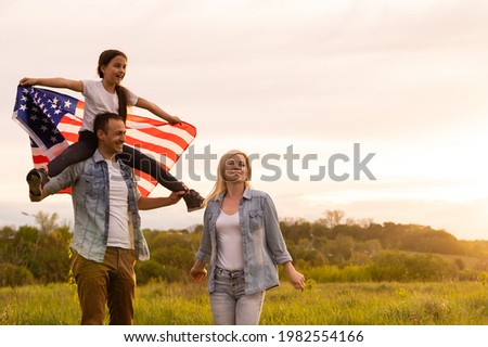 Young parents with their daughter holding American flag in countryside at sunset. Independence Day celebration