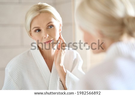 Headshot of gorgeous mid age adult 50 years old blonde woman standing in bathroom after shower touching face, looking at reflection in mirror doing morning beauty routine. Older skin care concept. Royalty-Free Stock Photo #1982566892