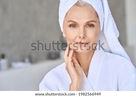 Happy smiling attractive senior older woman wearing bathrobe and white towel touching face looking at camera at bath room. Advertising of skin care spa procedures concept. Closeup portrait. Royalty-Free Stock Photo #1982566949