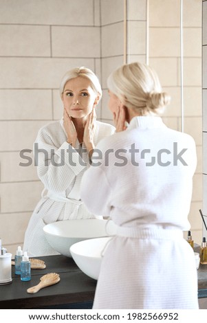 Attractive beautiful mid age adult 50 years old blonde woman standing in bathroom of SPA hotel touching face, looking at reflection in mirror doing daily morning beauty routine. Skin care concept. Royalty-Free Stock Photo #1982566952