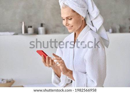 Portrait of happy smiling gorgeous middle aged lady wearing bathrobe and turban towel on head using mobile beauty apps holding smartphone. Antiage skin and hair care spa procedures concept. Royalty-Free Stock Photo #1982566958