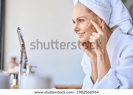 Gorgeous mid age older adult 50 years old blonde woman wears bathrobe in bathroom applying nourishing antiage face skin care cream treatment, looking at mirror doing daily morning beauty routine. Royalty-Free Stock Photo #1982566964