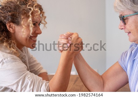 Family challenge, curly blonde daughter and senior mother joking arm wrestling on the table at home face to face