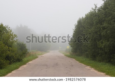 A deserted country road leading into the distance into the fog. Cracked asphalt on an old road. Trees and road signs on the roadside. Poor visibility in fog. Mystical mood.