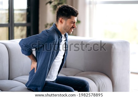 Sad upset young adult caucasian bearded man, in casual clothes, sitting on the sofa in the living room, massaging his back, suffering from back pain, suffering from arthritis, sciatica Royalty-Free Stock Photo #1982578895