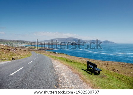 Small narrow road by the ocean, Achill Island, county Mayo, Ireland. Warm sunny day. Irish landscape. Blue clear cloudy sky. Small bench with spectacular view