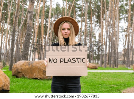 Portrait of caucasian young woman in hat holding cardboard with text NO PLASTIC outdoors. Nature background. Protester activist