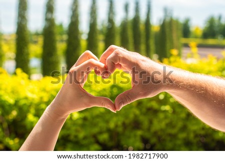 female and male hands in the shape of a heart, against the background of trees and a park. bright summer background. symbol of love and trust