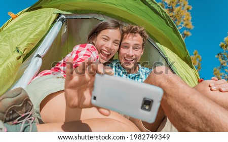 Camping selfie fun couple tourists holding phone taking self portrait photos in tent during summer vacation travel. Asian girl, Caucasian man laughing doing silly faces.