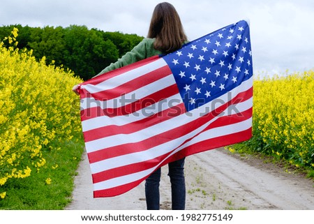 Girl holding waving on wind American national flag on a field road among yellow flowers. High quality photo