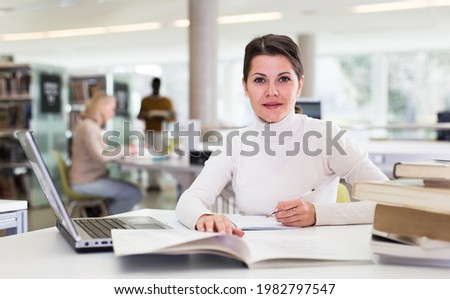 Portrait of smiling woman with laptop and book in public library. High quality photo