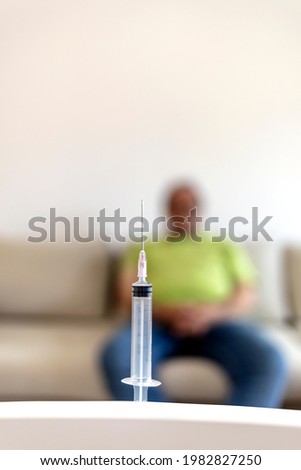 Photo of a syringe with a senior man blurred out in the background. Senior man looking at vaccine isolated on the white background. Happy elderly man is about to received dose of vaccine for COVID-19.