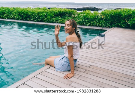 Smiling female tourist sitting on poolside with legs in water and waving hand saluting friend in tropical hotel near seaside