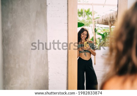Back view of young ethnic woman in stylish outfit standing against mirror and taking shot with mobile phone while resting on tropical resort during summer vacation