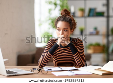 Stressed young afro american woman holding head in hands and feeling demotivated while sitting at her home office and working remotely on laptop. Depressed female student tired of onling learning Royalty-Free Stock Photo #1982859212