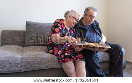 An elderly couple is sitting on the couch and looking at photos in a photo album.