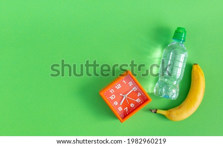 A bottle of still water, banana and an alarm clock on a green background. Time is morning. The concept of losing weight, healthy lifestyle, proper nutrition. World Obesity Day. Copy space.
