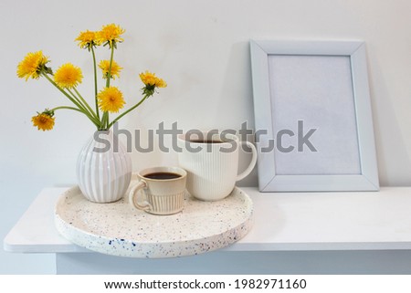 A bouquet of dandelions in a white fluted vase and two cups of different sizes with coffee on a white table and frame for picture. Breakfast. Copy space