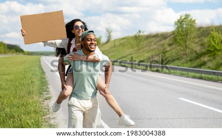 Wanderlust, autostop journey concept. Black guy and Caucasian woman hitchhiking on road with empty sign, travelling by autostop, having road trip, mockup for design. Panorama