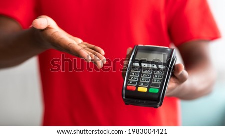 Digital Contactless Transaction. Cropped closeup view of African American man in red t-shirt uniform holding and showing POS machine, selective focus on payment dataphone terminal, blurred background Royalty-Free Stock Photo #1983004421