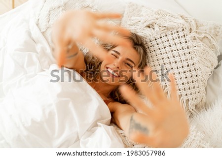Top view portrait of happy cheerful  woman lying in white cozy bed wake up in the morning, pulls  hands forward. Warm colors, boho interior. 