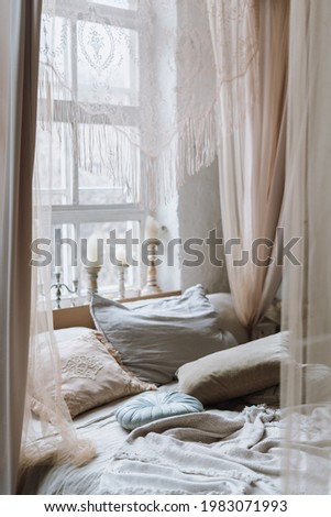 Vertical shot of cozy bed with various pillows and open baldachin, with light coming from big white vintage window with many different candles on windowsill. Empty bedroom in morning