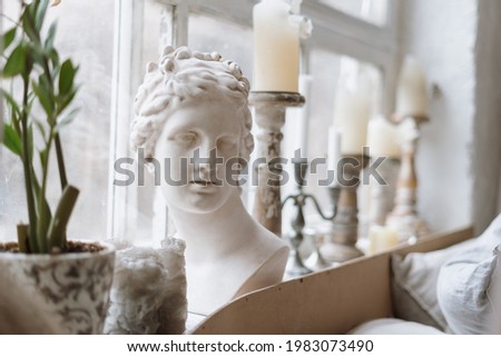 Close up shot of gypsum venus head on windowsill with antique candlesticks and green potted plant, blurred background with light coming from window indoors of cozy bedroom in morning