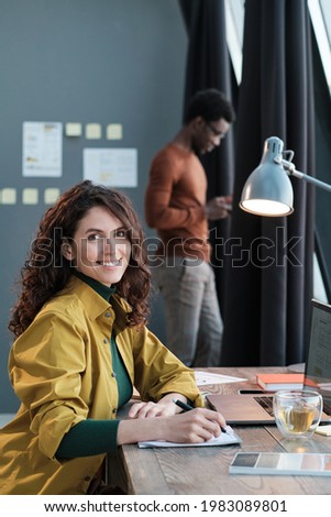 Portrait of young businesswoman smiling at camera while working at the table with her colleague in the background