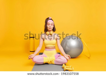 Calm relaxed young Asian woman meditates indoor sits in lotus position on karemat surrounded by sport equipment wears headphones around neck cropped top and leggings isolated on yellow wall.