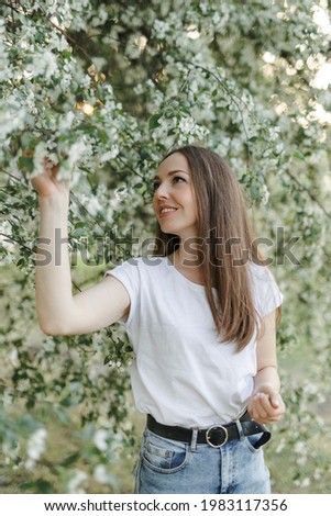Caucasian girl with brown hair in blue jeans and a white T-shirt on the background of an apple tree in bloom. Sunset. The concept of spring flowering. Apple tree in bloom.
