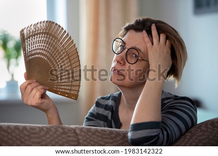 Exhausted middle aged woman waving her fan, suffering from menopausal symptoms, experiencing hot flush.  Royalty-Free Stock Photo #1983142322